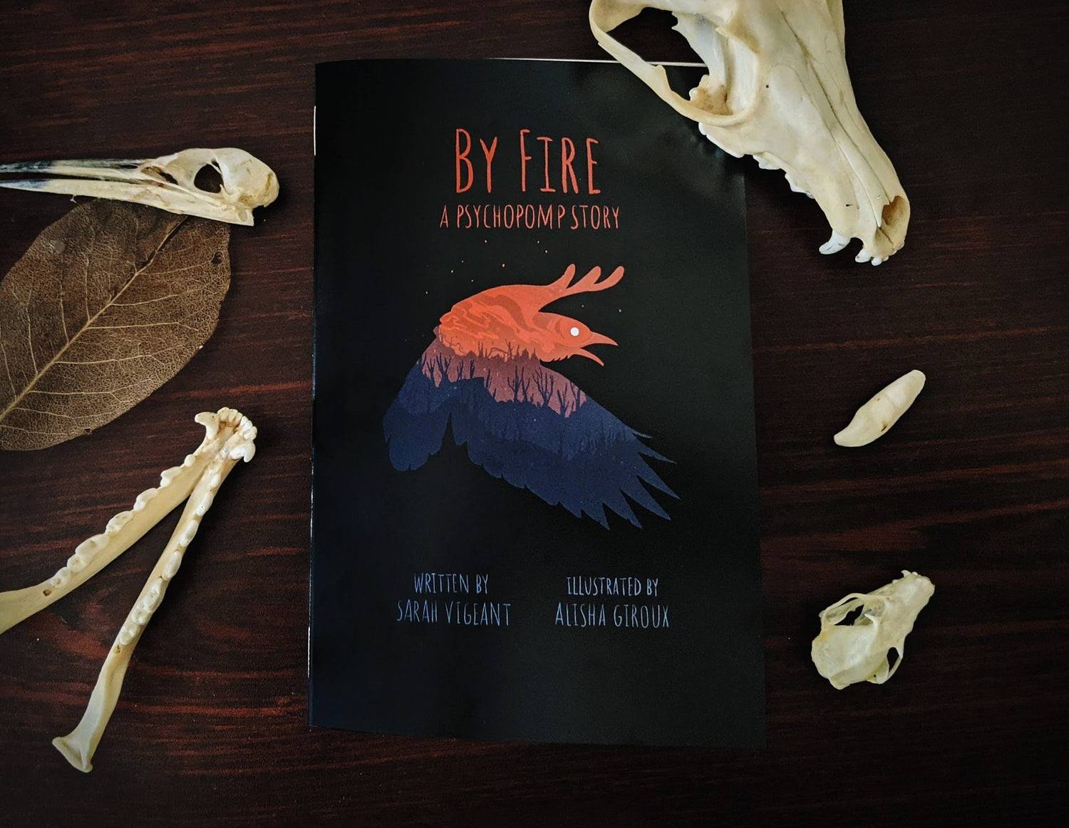 A comic book with a flying crow on the cover. The title reads 'By Fire'. The book lies on a dark table surrounded by animal skulls.