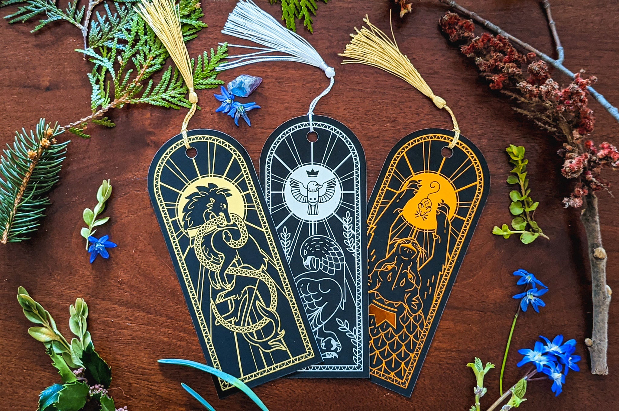 top down photo of three bookmarks, on a wooden background, surrounded by leaves and blue flowers. The bookmarks are all designed with metallic foil on black. Left to right, a golden hound with a snake in its mouth. The second is silver with a small crowned bird at the top, and a defeated eagle below it. The final is copper, depicting a mouse between the paws of a massive crowned lion.